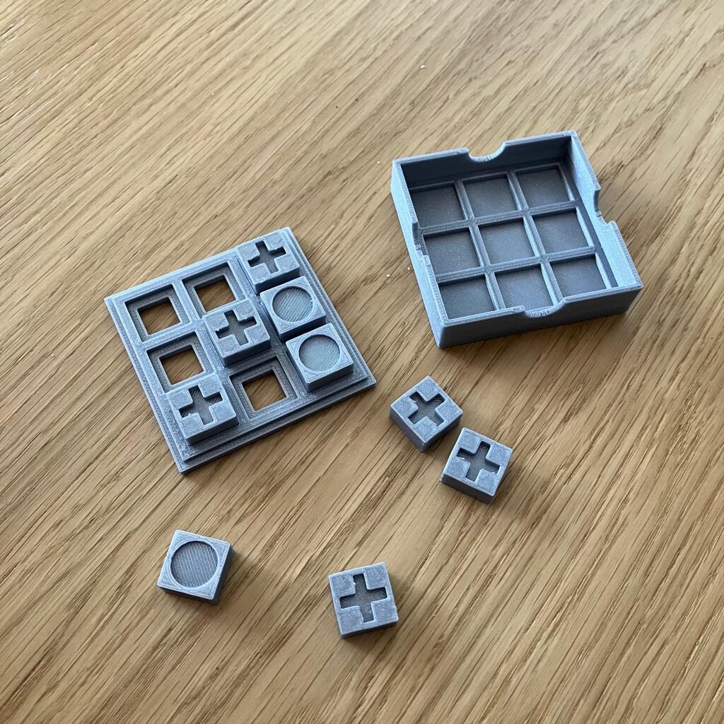 Kółko i krzyżyk - stół, gra leży w dwóch częściach, obok rozsypane cztery kostki: 3 z krzyżykiem, jedna z kółkiem. Na planszy ułożona skośna linia z trzech krzyżyków