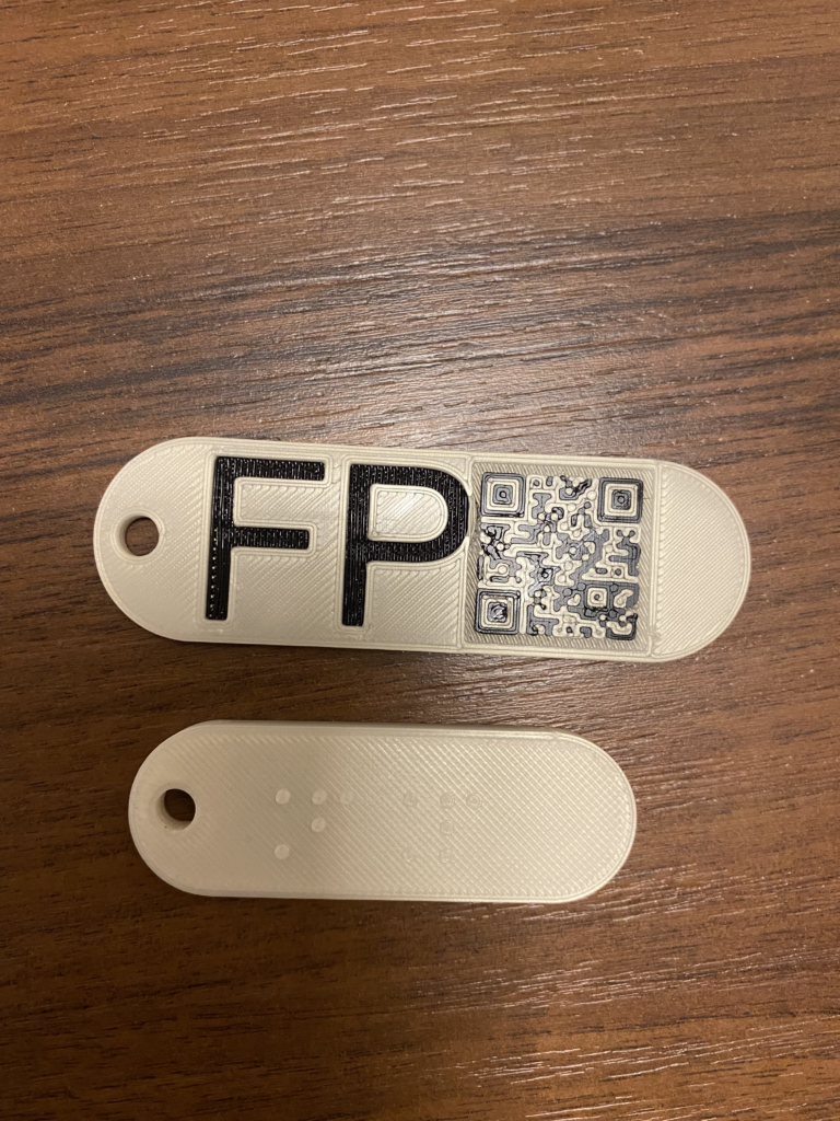 Dwa białe breloczki: na górnej powierzchni jednego widnieją brajlowskie litery FP, na drugiego czarny napis FP i kod QR.