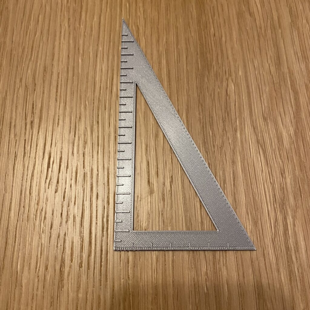 Srebrna ekierka prostokątna. Na krótszej przyprostokątnej znajduje się linijka z przedziałką co milimetr, na dłuższej, co pół centymetra.