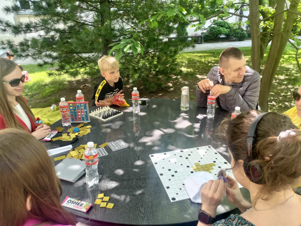 Pięć osób, wśród nich Julita i Monia, siedzi przy stole z grami. Widać scrabble, czwórki, kółko i krzyżyk, kostkę oraz domino.