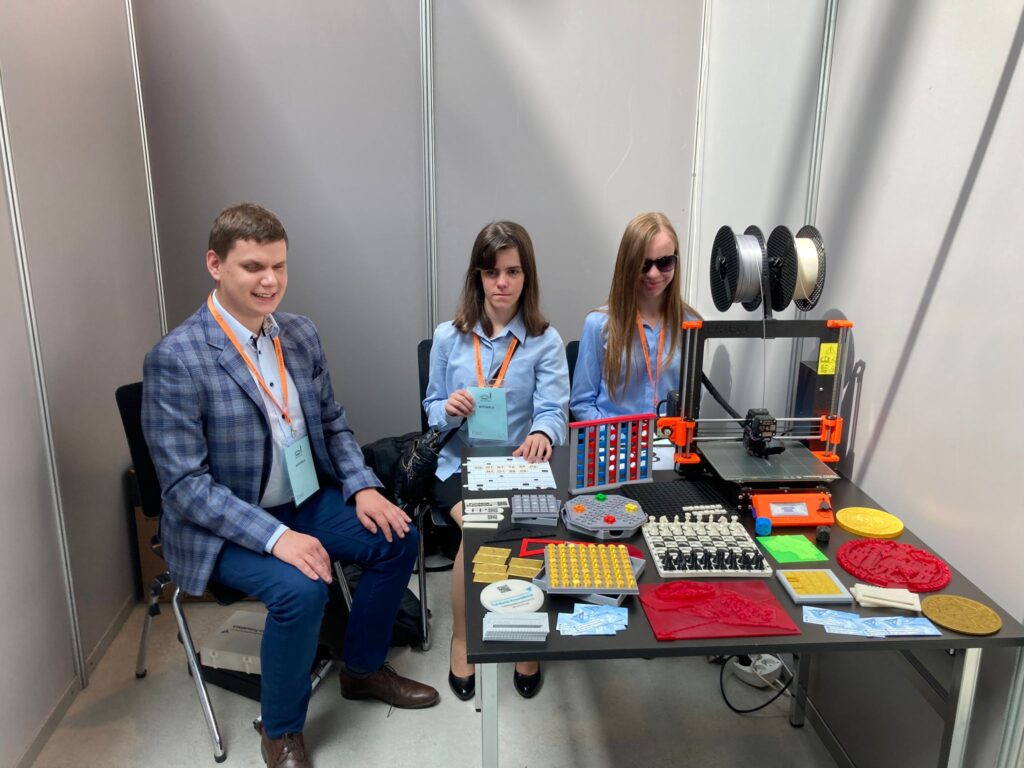 Dawid, Maja i Monika przy stanowisku z dalszej perspektywy. Widać cały stół i drukarkę. Maja trzyma w ręce plakietkę wystawcy.