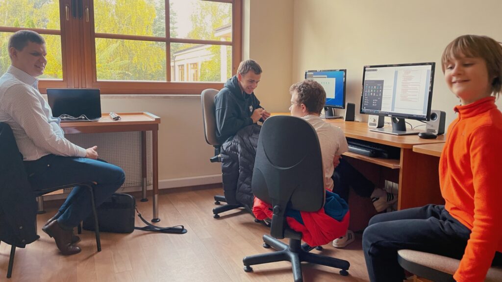 W pracowni informatycznej Dawid i troje uczniów przy komputerach twarzami do obiektywu, uczniowie wyglądają na ewidentnie zadowolonych ze spotkania samego twórcy Eltena. ;) Dawid ubrany jest w eleganckie granatowe spodnie, białą koszulę z szarym wzorem i brązowe buty.