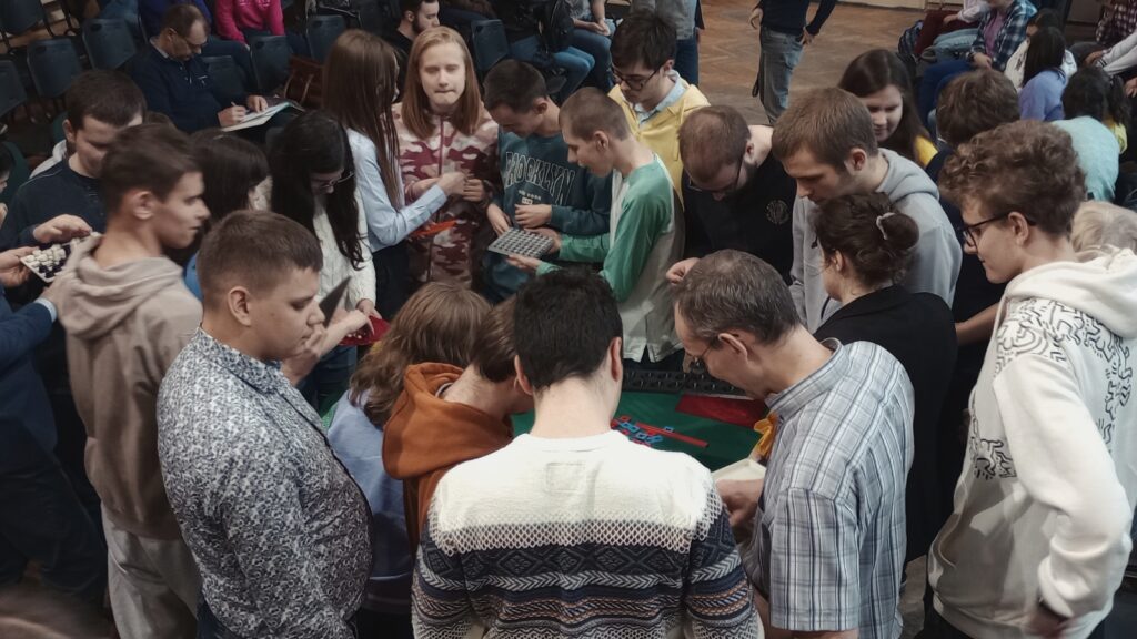 Widok z góry na wielu uczniów wokół stołu z wydrukami; w tłumie można rozpoznać Dawida i Julitę, którzy rozmawiają z uczniami